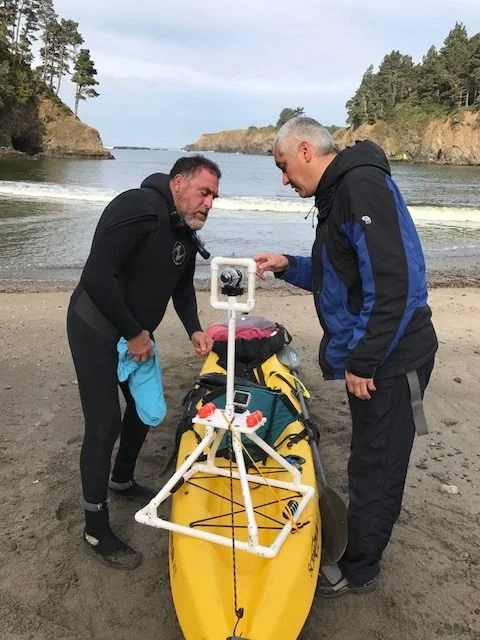 Two men on a beach mounting a camera to a white frame on a yellow sea kayak, a cove behind them
