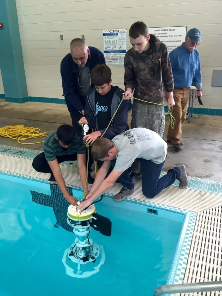 Students and instructors kneeling at the edge of an indoor pool, lowering a tethered underwater robot into the water